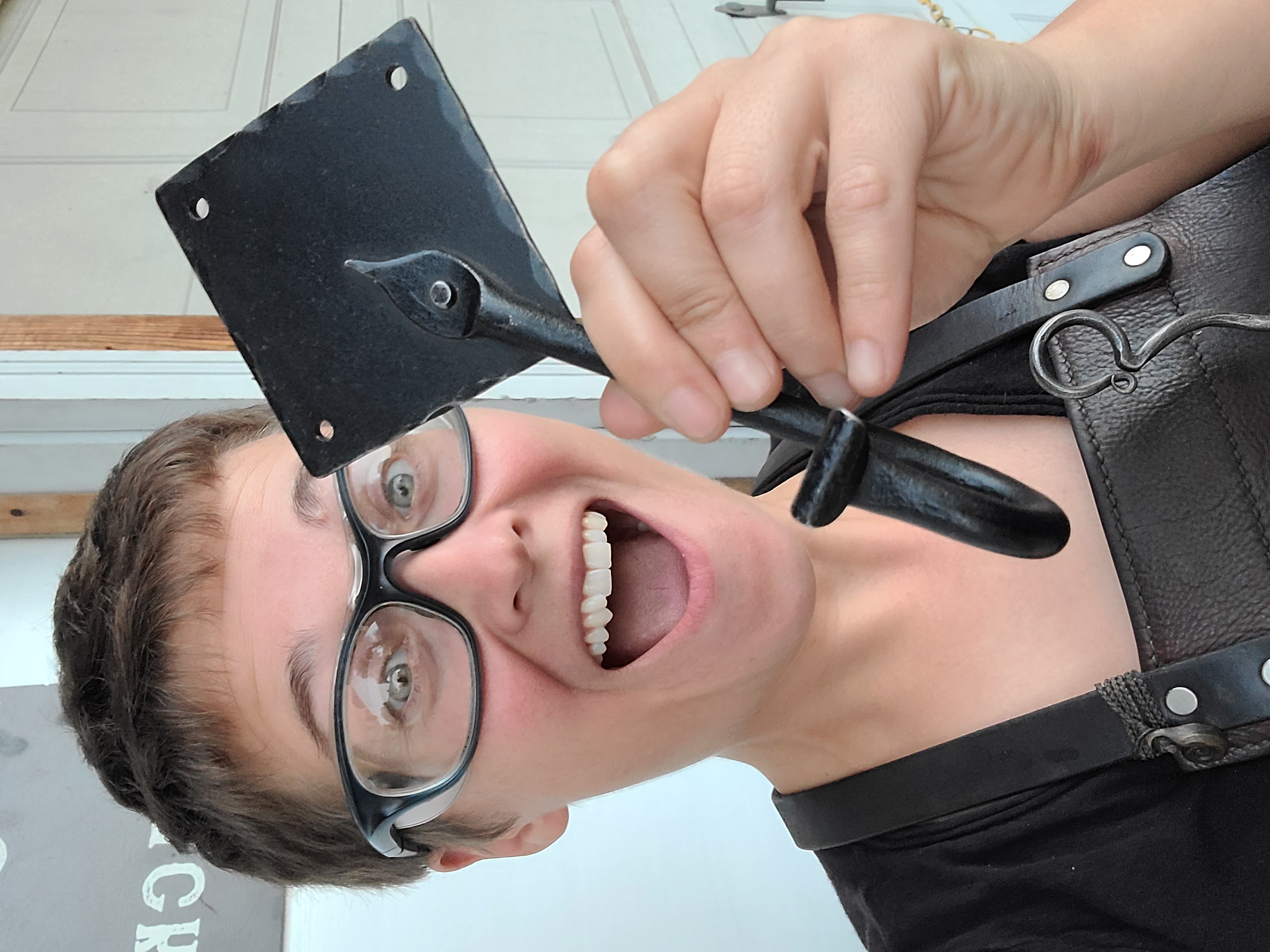 Ms Caitlin holds up a black steel J-hook with a diamond backing plate. She is wearing a leather apron with a hand-forged pointer and she looks excited. 