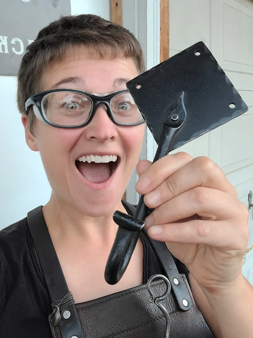 Ms Caitlin holds up a black steel J-hook with a diamond backing plate. She is wearing a leather apron with a hand-forged pointer and she looks excited. 