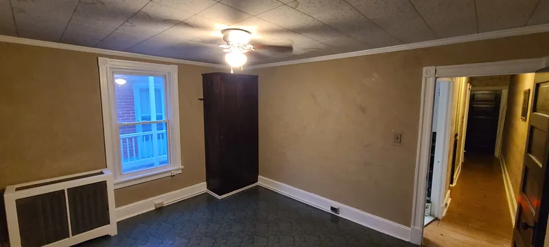 View from back bedroom down the hall. Taupe plaster walls, white trim, dark furniture, white and bronze radiator cover, ceiling fan, green vinyl flooring. 