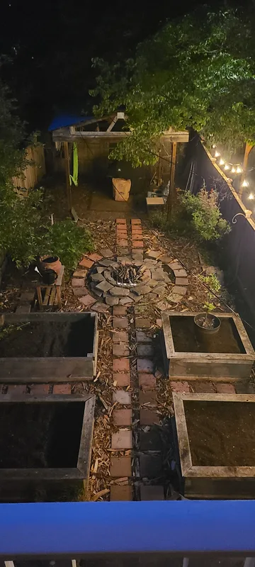 View of the back yard from the upstairs porch. 4 raised beds, red pavers around the garden beds, fire pit in the middle. Hammock shelter in the back. Fig tree to the left. Night time photo with the neighbor's string lights on. 