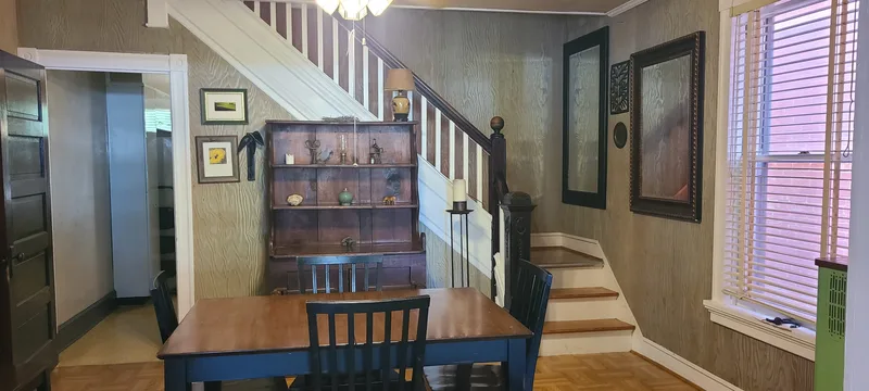 Dining room with view of kitchen to left, stairs that run across the back of the room from the bottom right. White trim, dark wood, light wood, paneling