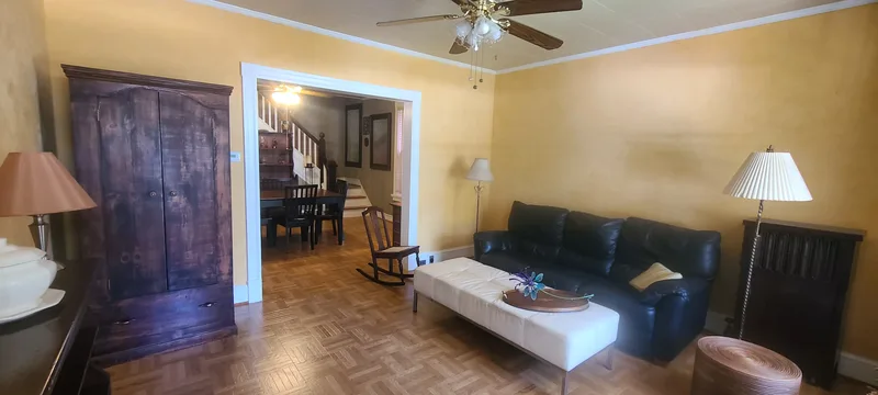 Living room to dining room view with a wider angle lens. Yellow walls, white trim, vinyl parquet floors, dark furniture, a bronze and glass ceiling fan